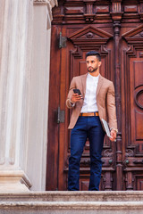 Young East Indian American Man with beard, wearing light brown blazer, white round collar shirt, blue pants, holding laptop computer, standing in front old style door way, texting on cell phone..