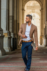 Man Street Fashion.Young East Indian American Businessman traveling, working in New York City, wearing brown blazer, blue jean, holding laptop computer, cell phone, walking on vintage narrow street..