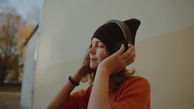 cute young girl listening music in headphones and dancing, urban style, stylish hipster teen in black hat listen music and smile infront of yellow wall, orange crazy street style