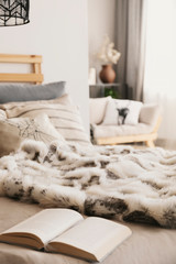 Bed with open book, pillows and fur coverlet in Scandi apartment interior in real photo with blurred background