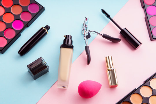 Top view picture of makeup products: lipstick and concealer palettes, foundation, mascara and eyelash curler on pink and blue background. Make-up artist kit concept