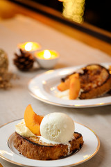 Delicious French Toast served with peach and ice cream on a ceramic dish. Decorated table with candles and pine cone, on a cloth background