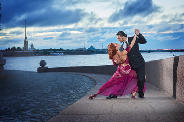 Young, beautiful and professional couple dancing tango on the quay of the river at daybreak in Saint-Petersburg on Peter and Paul Fortress and Neva river background - obrazy, fototapety, plakaty