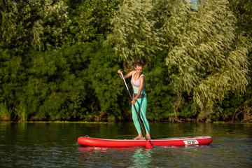 Young athletic woman doing fitness on a board with an oar on a lake.