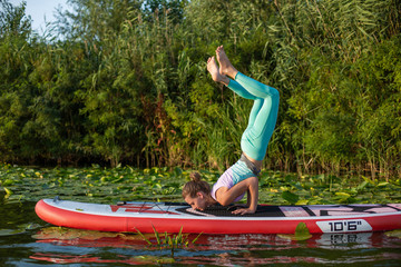 Photo of young woman doing hand stand on stand up paddle board. She wearing a leggings and top.