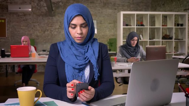 Serious face of amuslim female in blue beutiful hijab is scrolling her phone while sitting in modern studio, surrounded with other working arab girls