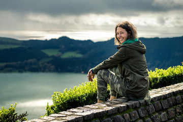 Portrait of young woman in khaki clothing sitting on the view point and smiling, looking at camera....