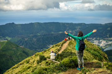 Fototapeta na wymiar Young woman in khaki clothing is standing on the most popular place on Azores islands - Miradouro da Boca do Inferno. Backside view and raised up hands. Portugal, Sao Miguel.