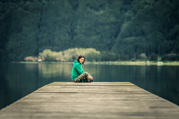 Portrait of young woman in green sweatshirt sitting on the edge of pier. There is volcanic lake on the background. Peaceful atmosphere. Smiling and looking at camera. Azores islands, Portugal
