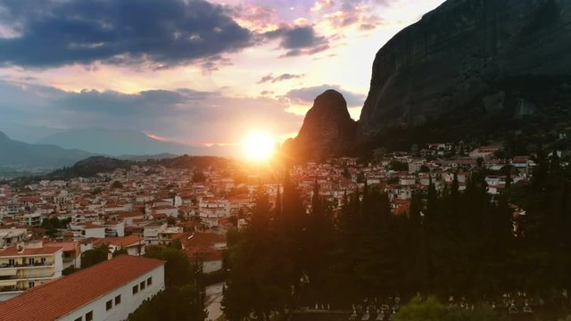 Aerial shot taking off over old European city Kalambaka or Kalampaka or Kalabaka with small white houses and orange roofs great view of mountain Meteora at sunset amazing colored sky and yellow sun