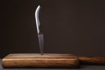 Knife stuck in cutting board on dark background. Wood cutting board and kitchen knife against dark brown wall with copy space. - Powered by Adobe