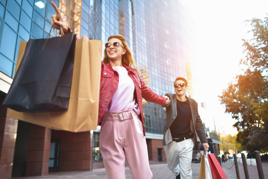 Front View Of A Casual Couple Of Shoppers Running In The Street Towards Camera Holding Colorful Shopping Bags.