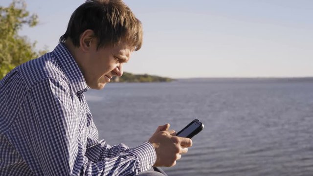 Man in a Checkered Shirt Looks at a Smartphone Sitting on the River Bank