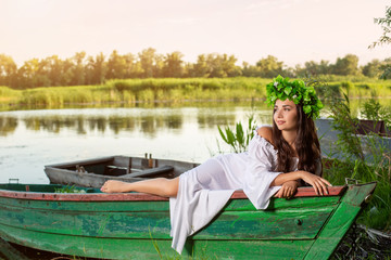 The nymph with long dark hair in a white vintage dress sitting in a boat in the middle of the river.