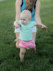 Mother is helping her baby boy taking his first steps.A small girl boy with dark eyes in a white shirt and denim shorts, trying to take the first steps with the help of his mother