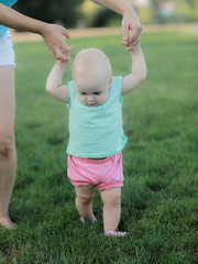 Mother is helping her baby boy taking his first steps.A small girl boy with dark eyes in a white shirt and denim shorts, trying to take the first steps with the help of his mother