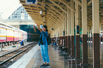 Asian woman traveler used smart phone take photos while wait and check train schedule on the platform of the railway station - travel and transportation concept