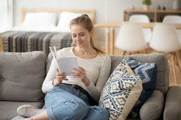 Obraz premium Young attractive positive woman spend free time on weekend alone sitting on couch in living room holding a book at modern cosy home. Millennial smiling girl enjoy reading love story novel or chicklit