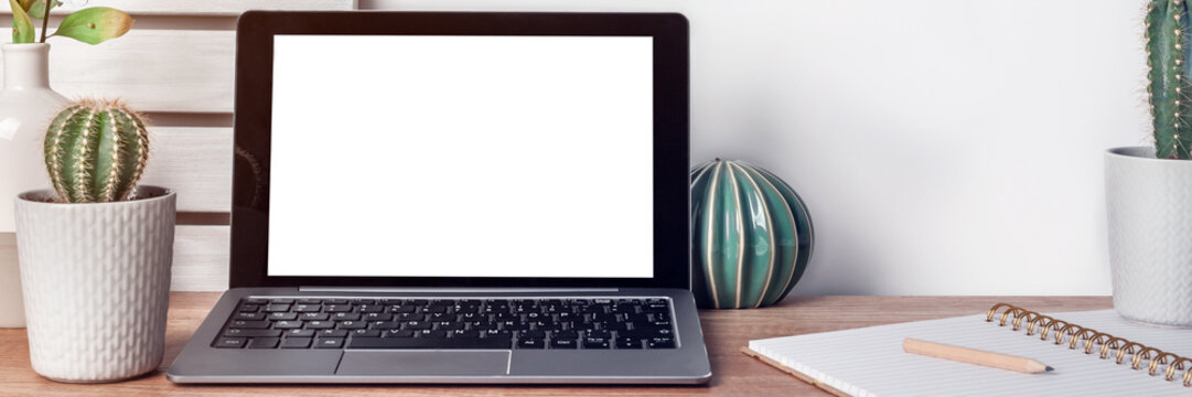 Panoramic photo of hipster wooden desk with laptop mockup, cactus in a gray pot, a vase, ceramic ornament, open notebook and boards on the wall