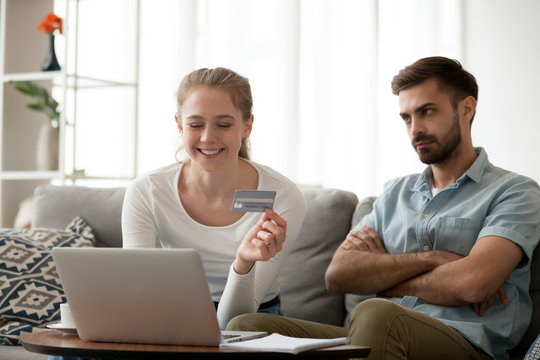 Unsatisfied husband looking at smiling wife, girl holding credit card choose goods on internet online making shopping wasting money using laptop sitting at home. Extra expenses and big spender concept
