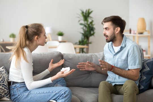 Unhappy young millennial married couple sitting on couch in living room at home and quarrelling. Husband and wife emotionally arguing talking cheating. Break up and problems in relationships concept