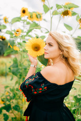 Naklejka premium Young amazing beautiful blond girl in black dress with ethnic pattern lifestyle portrait. Country fashion. Cute odd happy cheerful female in traditional slavonic clothes posing in sunflowers outdoor.