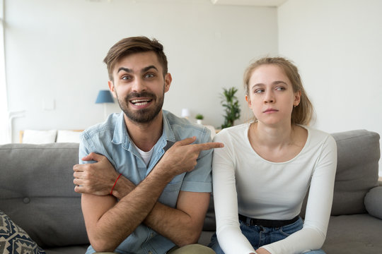 Millennial married couple sitting together on couch in living room at home. Boyfriend and girlfriend having video call funny male pointing on dissatisfied female complains talking with friends