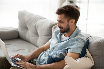 Fototapeta premium Young millennial positive man sitting on couch in living room with laptop alone on weekend at home. Handsome male using notebook have fun looking recreational video or freelancer working online indoor