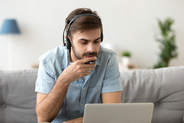 Man sitting on couch at home studying using computer and headset looking at device screen listening...