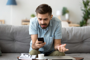 Confused millennial man sitting alone on couch in living room at home looking at smartphone screen...