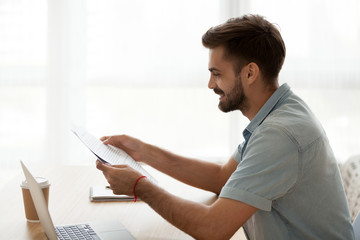 Happy satisfied millennial man sitting at the desk holding letter paper about recruited hired on a good post. Student reading pleasant news about scholarship or examination test passed successfully