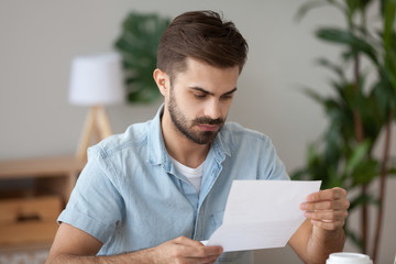 Frustrated millennial man sitting at the desk in office holding reading notification about debt or...