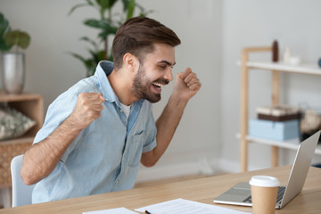 Millennial man sitting at the desk looking at computer device screen celebrating win feels happy...