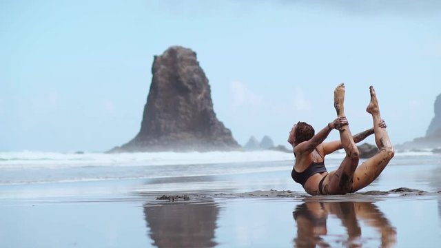 A beautiful woman in a swimsuit performs an exercise bridge on the beach with black volcanic sand surrounded by mountains and ocean. Gymnastics and yoga