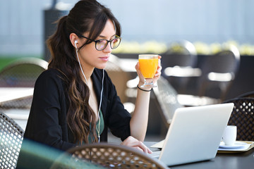 Beautiful young woman drinking orange juice while working with her laptop in a coffee shop.