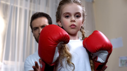 Pretty kid in boxing gloves practicing punches with father behind, courage