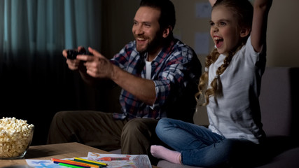 Excited father and daughter playing video game, female kid raising hands, winner