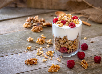 White yogurt with muesli in glass bowl with pieces raspberries on top on natural wooden desk.