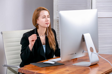 Portrait of happy young successful businesswoman at office. She is sitting at the table, holding her glasses and looking thoughtfully at display. Black Friday or Cyber Monday. Selecting items