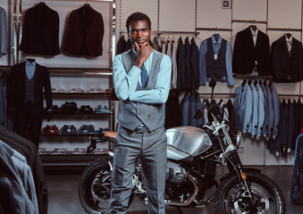 Elegantly dressed African American young man posing near retro sports motorbike at the men's clothing store.