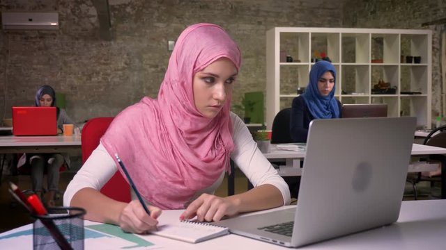 Nice working arab female in pink hijab and islamic clothes is writing down notes and looking at her laptop precisely while sitting in front of other muslim women, office vibes
