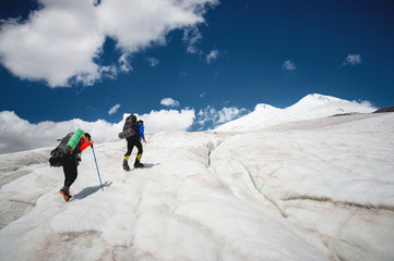 Two tourists, a man and a woman with backpacks and crampons on their feet walk along the glacier against the background of the mountain Elbrus and the sky and clouds. Back view