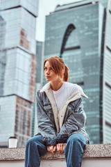 Stylish redhead hipster girl with tattoo on her face wearing denim jacket sitting in front of skyscrapers in Moskow city at cloudy morning.