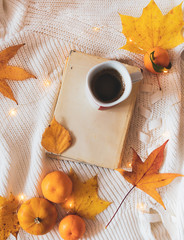Flat lay or top view of a book with old covers, cup of coffee, pumpkin, mandarin oranges and golden leaves and lights on white woolen sweater. Autumn or winter morning coffee or breakfast background. 