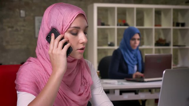 Confident nice muslim girl is focused having phone talk all in her minds, sitting in brick office with other arabic girls on their desktops, modern lifestyle