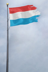Fototapeta na wymiar Worn and tattered Luxembourg flag blowing in the wind on a cloudy day