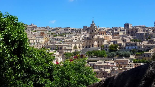 Modica, Sicily - Italy landscape with the catholic church (Cathedral of San Giorgio) front view. Old modica houses around the church. Leaves of the top of a tree sway in the foreground.