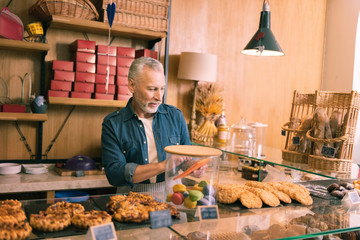 Selling cookies. Bearded grey-haired owner of bakery feeling joyful while selling cookies in the morning