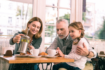 Pouring tea. Beaming blonde-haired grandmother pouring some tea for her girl while having breakfast in cafeteria