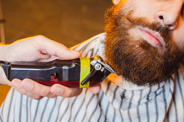 Barber cuts a beard with a trimmer to a young handsome guy with a mustache. Haircut of a beard in a...
